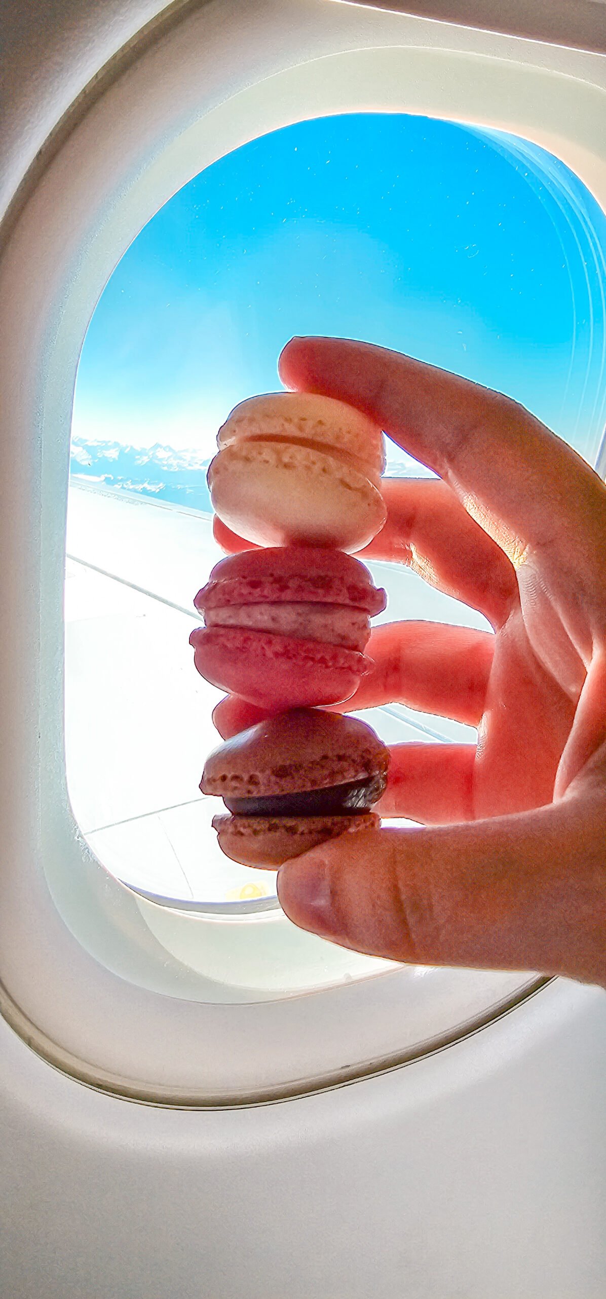 Guia de melhores restaurantes onde comer em Zurique 13 Aceitamos Viagens onde comer em zurique Uma janela de avião com uma mão segurando três macarons, um de chocolate, um de framboesa e um de baunilha